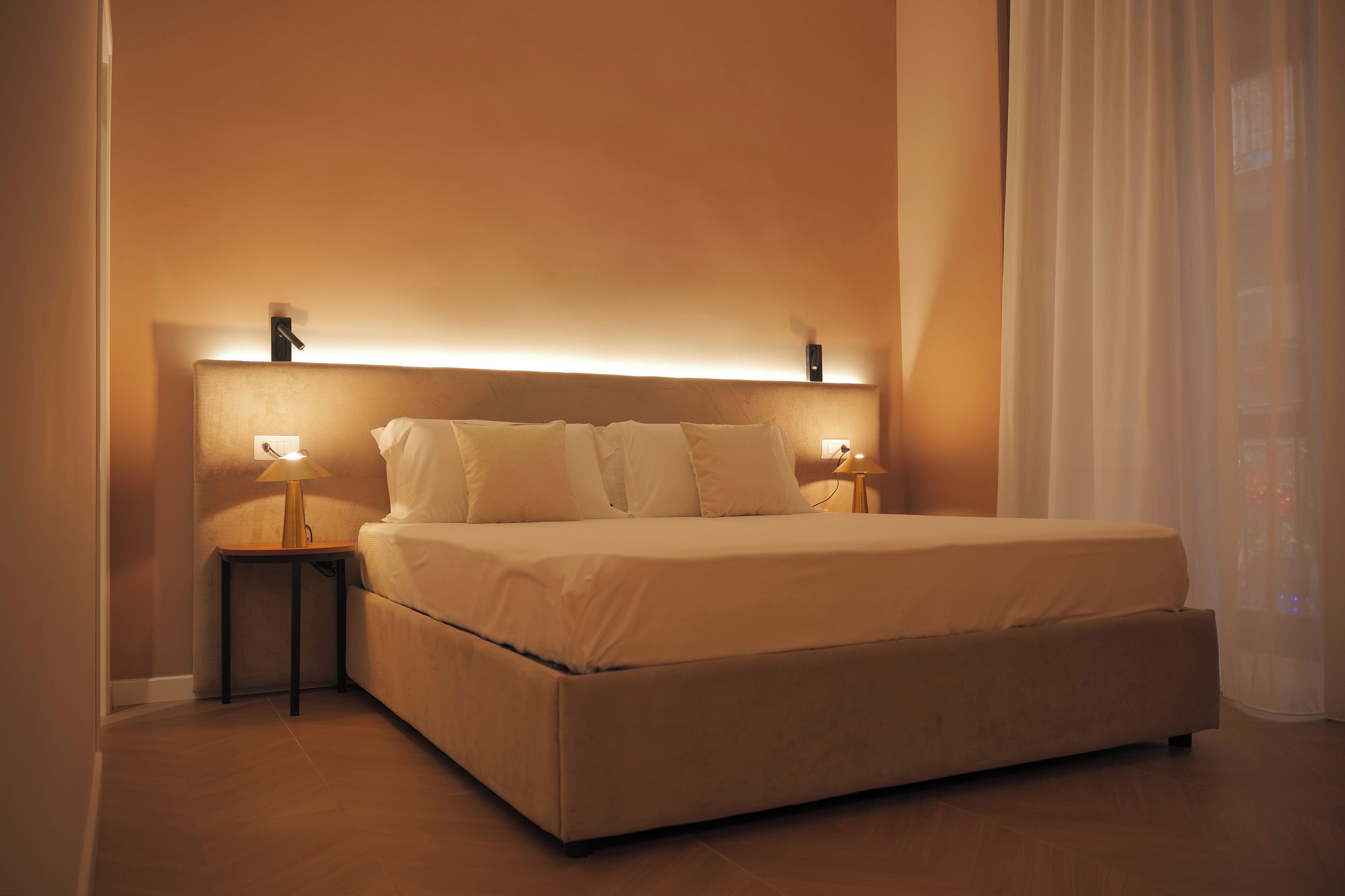 A neatly made bed with a headboard, two bedside tables with lamps, and sheer curtains.