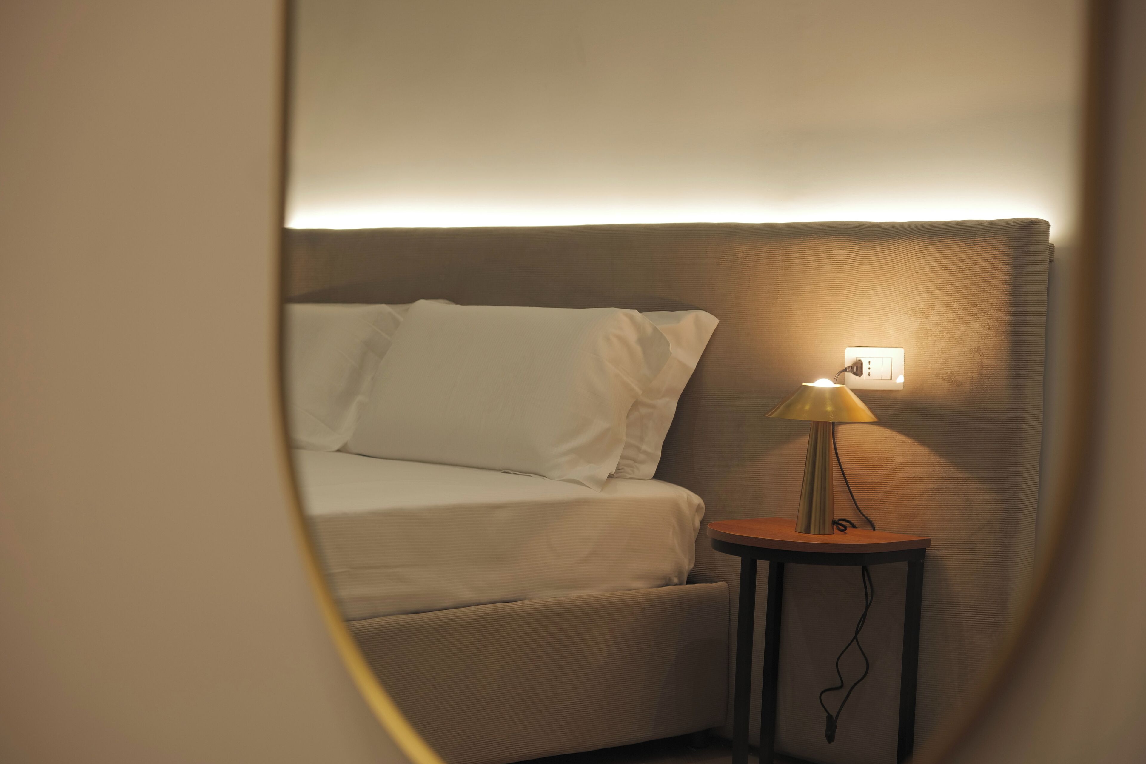 Un lit avec du linge de lit blanc, une petite table ronde avec une lampe et une prise de courant au mur.