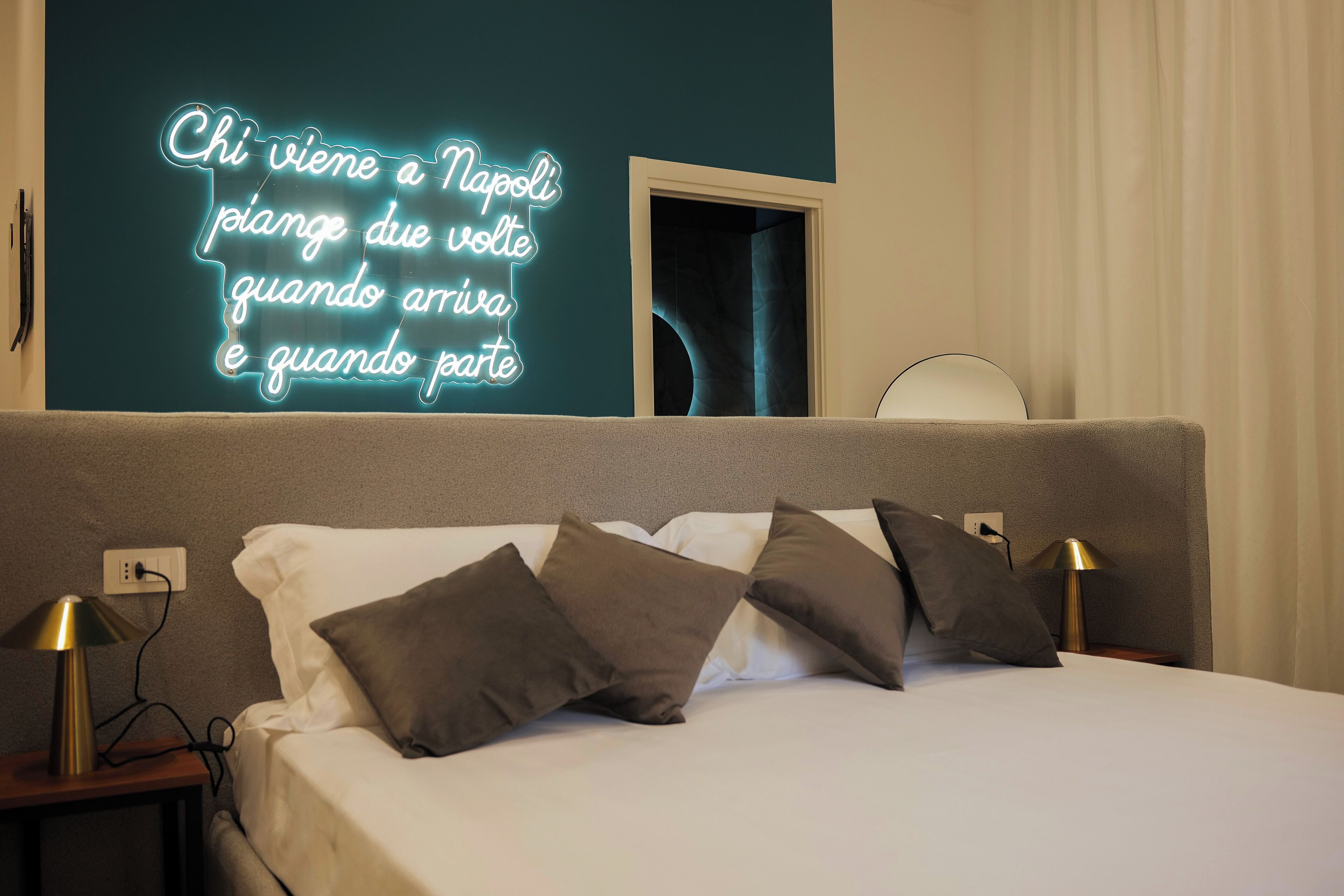 A hotel room with a bed, pillows, bedside lamps, a neon sign, and a window with curtains.