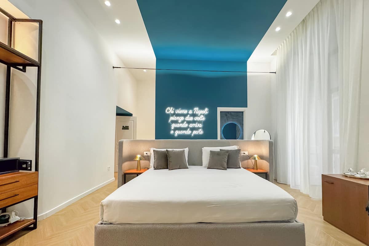 A modern bedroom with a large bed, bedside tables, a neon sign, and a view of the city.