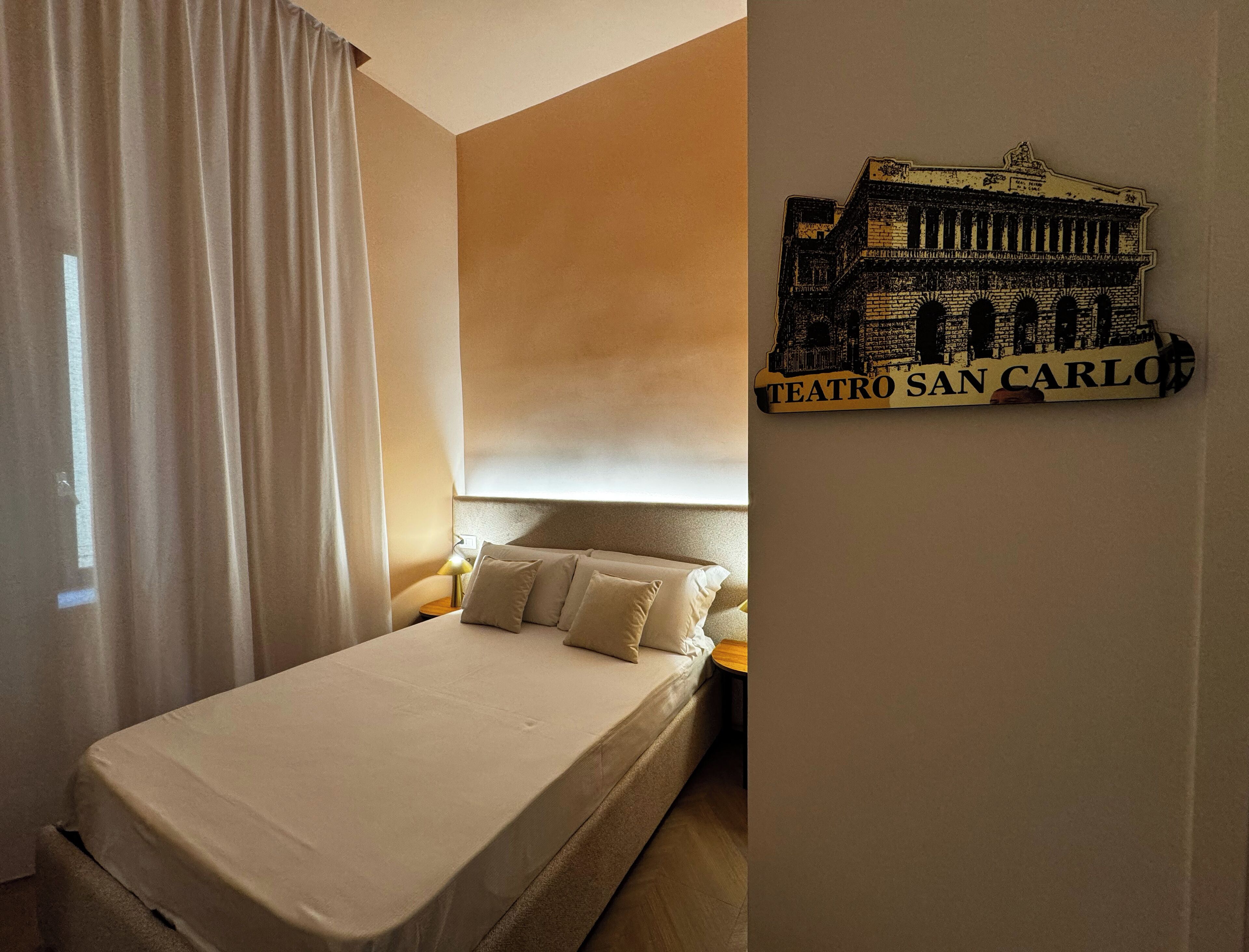A hotel room with a bed, a nightstand, a lamp, and a wall-mounted sign reading 'TEATRO SAN CARLO'.