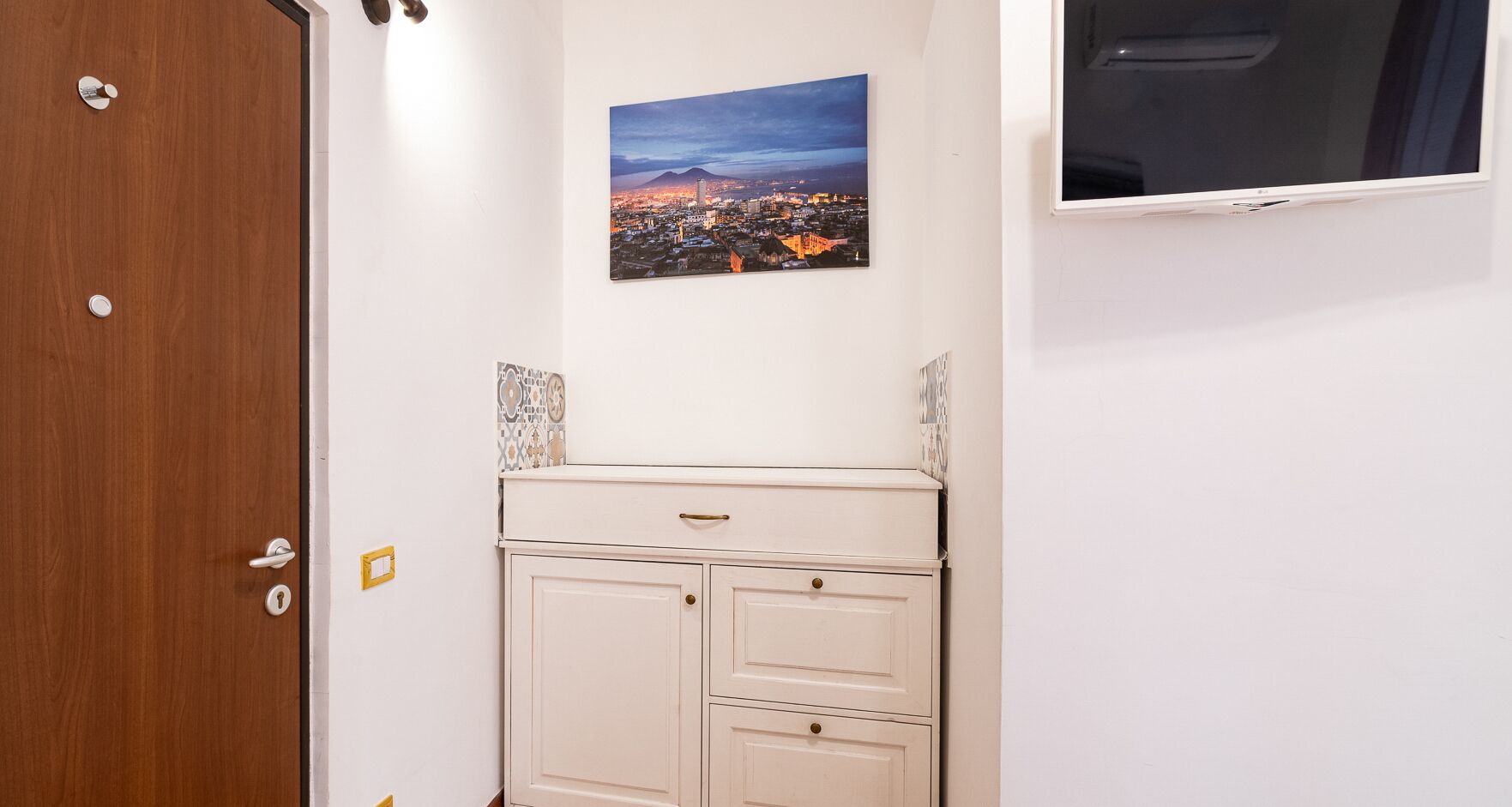A room with a wooden door, a white cabinet with drawers, a framed cityscape picture on the wall, and a flat-screen TV mounted above the cabinet.