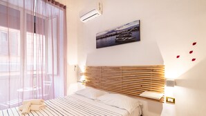 A bedroom with a bed, a window with sheer curtains, an air conditioning unit, a wall-mounted picture, and a bedside table with a lamp. - Domus Mattei al Duomo By DomusExtra (Naples)