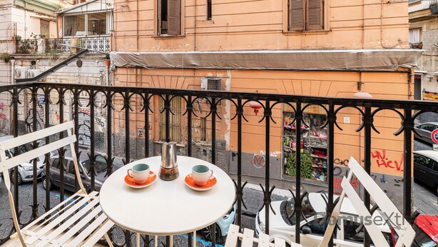 A balcony with a table and chairs, a railing, and a view of a street with parked cars and buildings. - Domus Mattei al Duomo By DomusExtra (Naples)