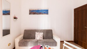 A living room with a grey sofa, a small table with a pink cloth, a framed picture of a coastal town, and a wooden door. - Domus Mattei al Duomo By DomusExtra (Naples)