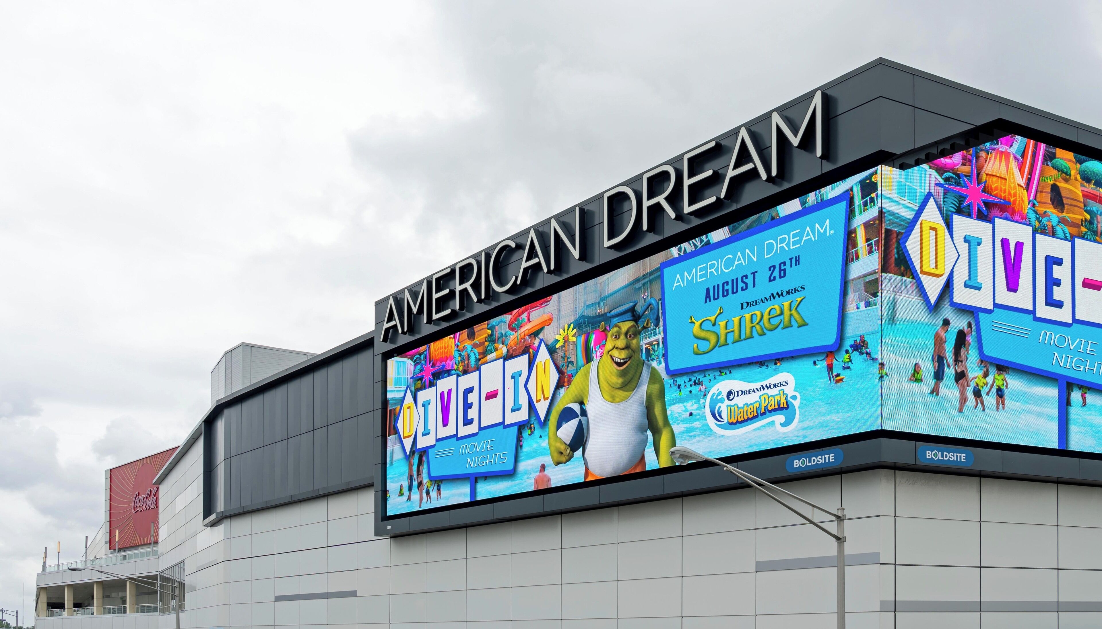 Ein Gebäude mit einer großen digitalen Werbetafel, die 'American Dream' und Filmabende von 'Shrek' bewirbt.