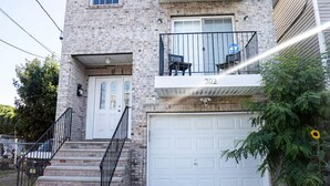 A two-story brick house with a white garage door, a balcony with a metal railing, and a small front porch with steps.