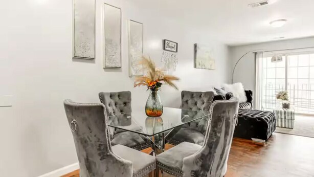 A dining area with a glass table surrounded by four grey upholstered chairs, a vase with dried flowers, and framed artwork on the wall.