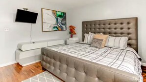 A modern bedroom with a tufted headboard, a large bed with a checkered bedspread, a flat-screen TV mounted on the wall, and a minimalist bedside table.