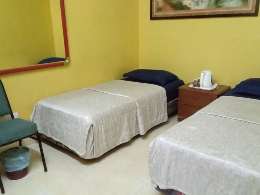 A room with two beds, a chair, a small table with a kettle, and a trash bin.