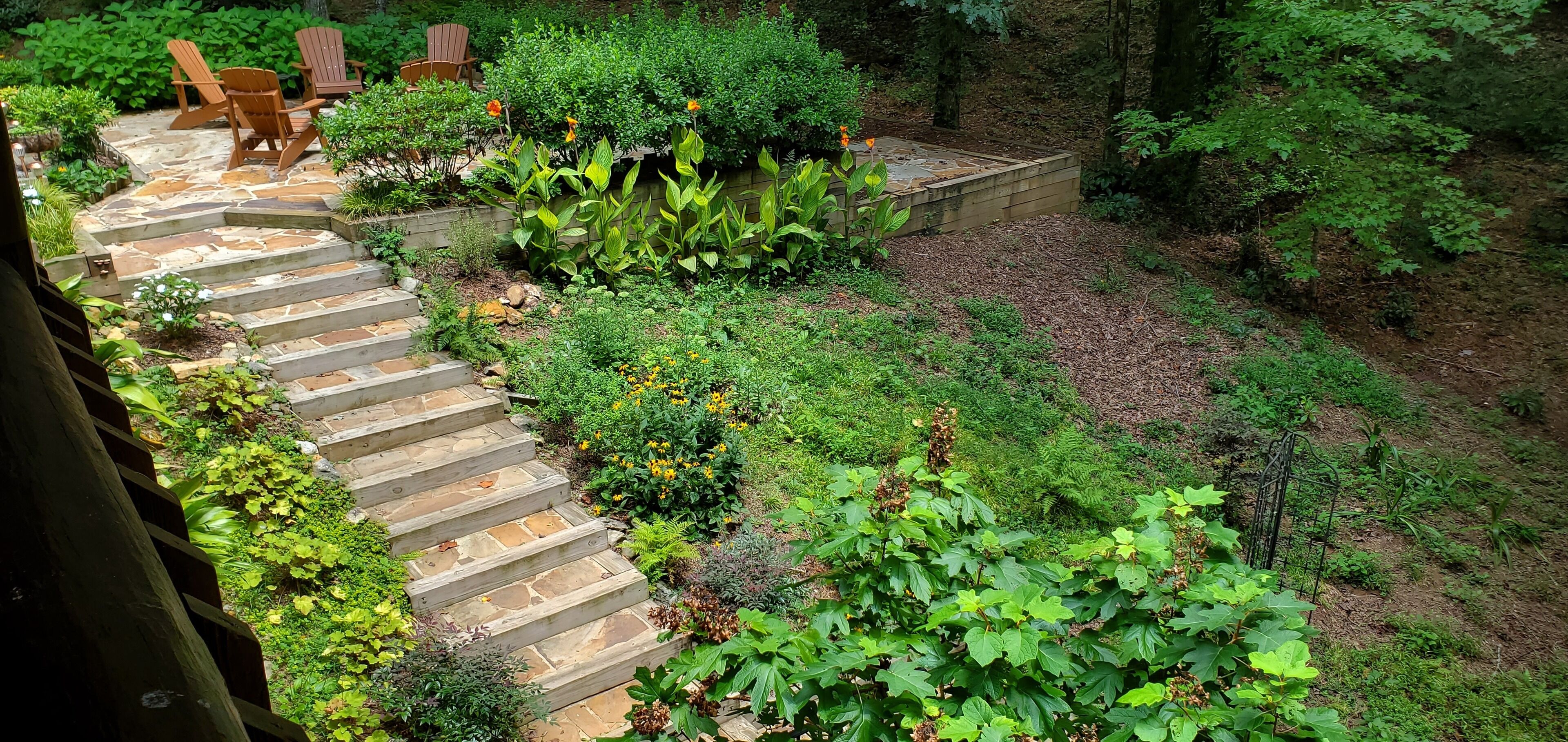 A stone staircase leading to a wooden deck with Adirondack chairs, surrounded by lush greenery and a variety of plants.