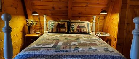 A bedroom with a wooden ceiling, a bed with a quilt, two bedside lamps, a nightstand with a vase, and a dresser.