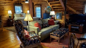 A cozy log cabin interior with a wooden floor, a sofa, a chair, a television, and a lamp.
