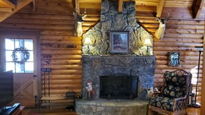 A log cabin interior with a stone fireplace, wooden walls, a television, and a cozy seating area.