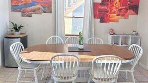 Een eetgedeelte met een houten tafel en witte stoelen, een koelkast, een cactus en kunst aan de wanden.