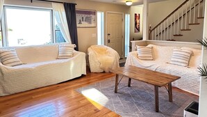 Een ruime zithoek met een houten vloer, twee witte banken met gestreepte kussens, een houten salontafel en een trap naar een hoger niveau.