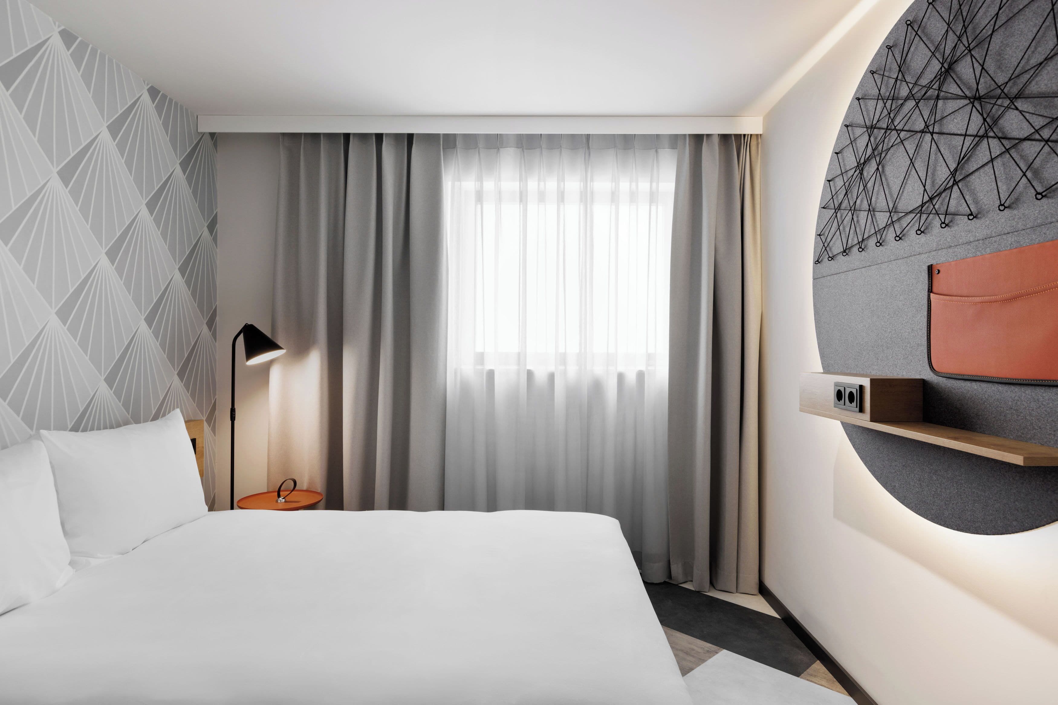 A modern hotel room with a large bed, a bedside table, a lamp, a window with sheer curtains, and a wall-mounted shelf with a leather bag.