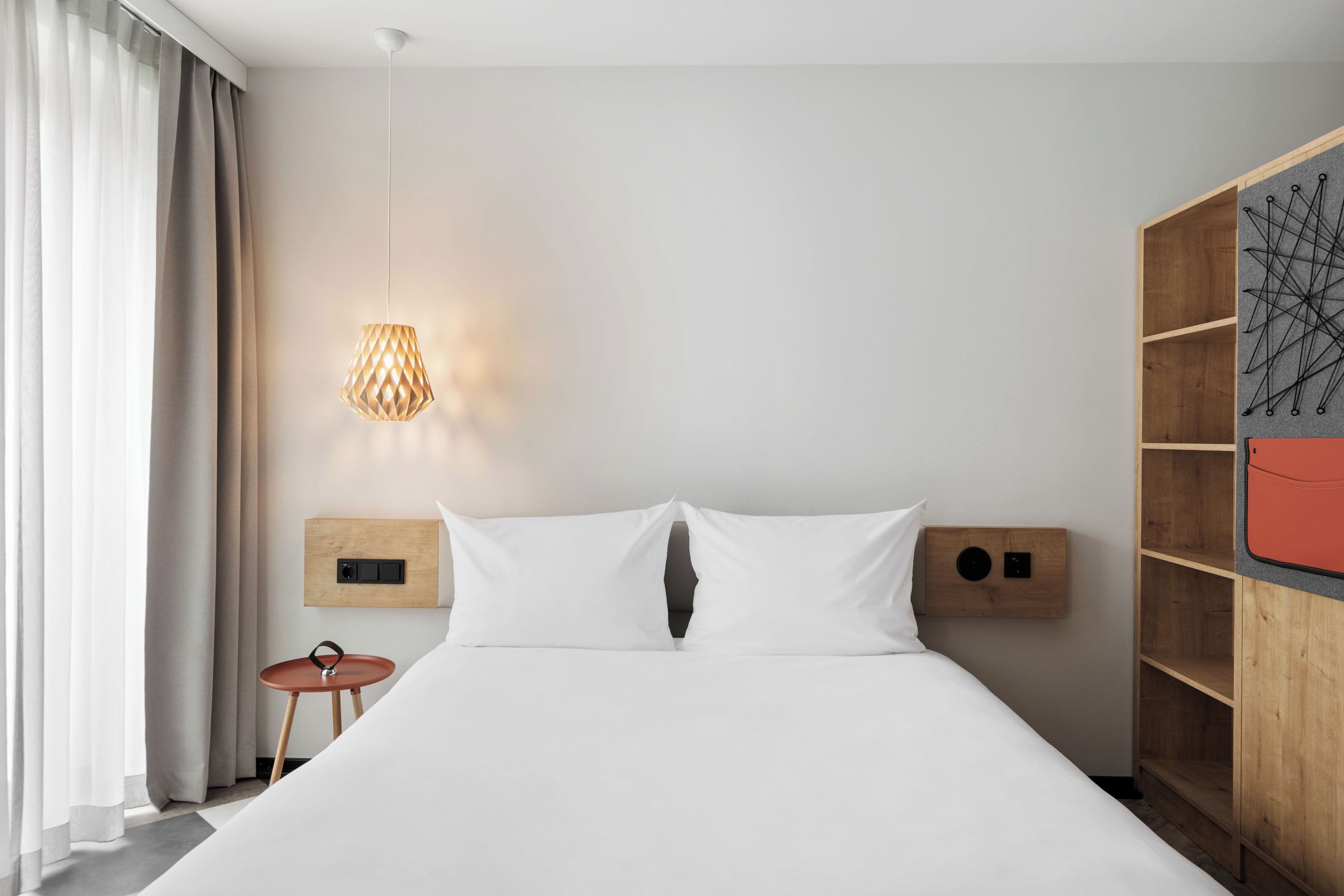Quarto de hotel moderno com cama, mesas de cabeceira, luminária na parede e guarda-roupa de madeira.