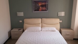 Una cama doble con un edredón blanco acolchado, dos mesitas de noche con lámparas, y un cuadro enmarcado con una vista urbana en la pared.