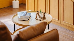 Um quarto aconchegante com mobiliário de madeira, uma pequena mesa redonda com livros e uma planta, e uma estante com revistas e uma moldura com uma fotografia.