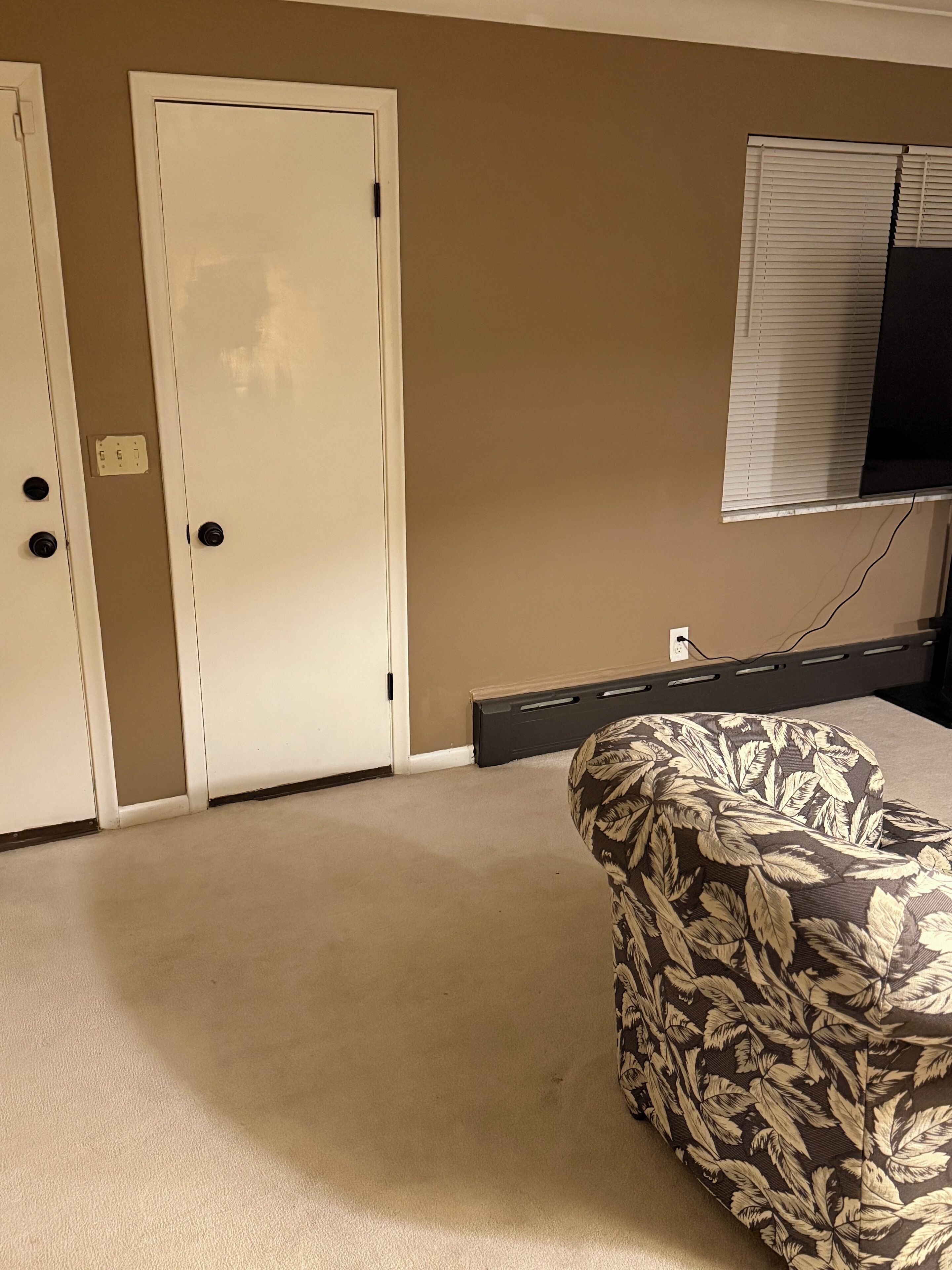 Una stanza con tappeto beige, una porta bianca, una TV a muro e una sedia a fantasia.