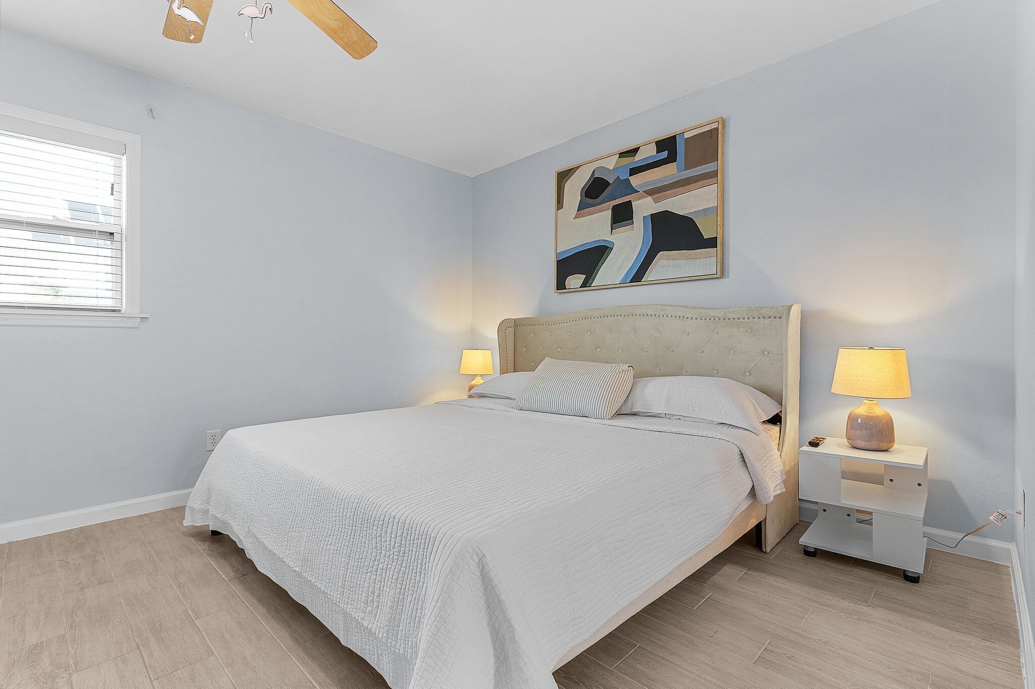 A bedroom with a bed, bedside lamps, a nightstand, a ceiling fan, and a wall-mounted abstract painting.
