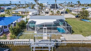 Une maison dotée d’un grand espace piscine entièrement vitré, d’une jetée en métal s’étendant sur une étendue d’eau, et entourée d’un quartier résidentiel planté de palmiers et comprenant d’autres maisons.