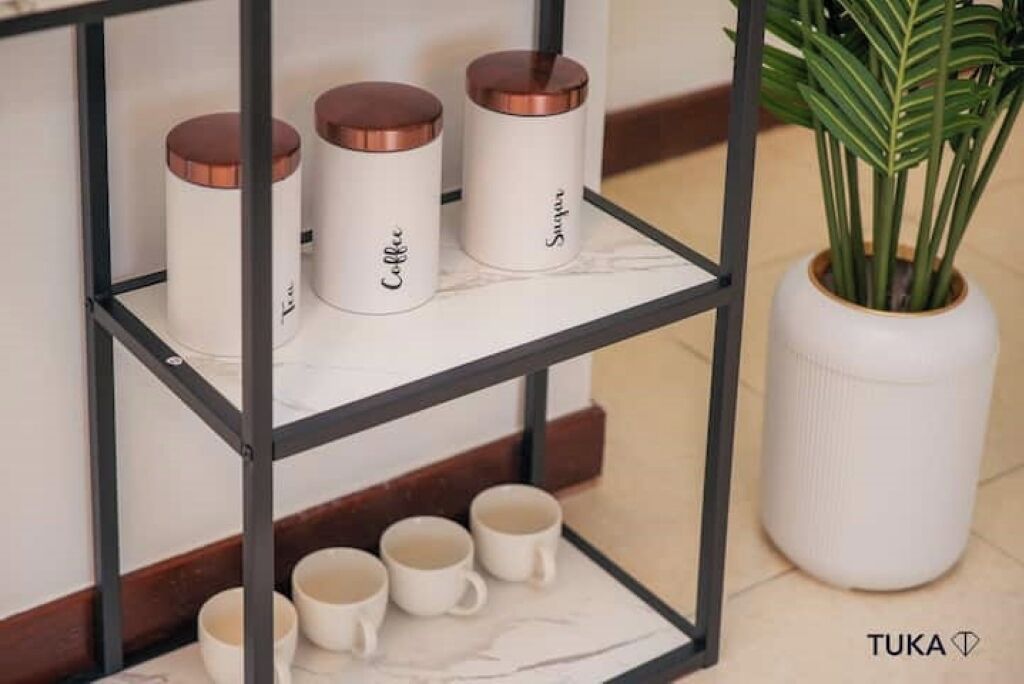 A shelf with three white containers labeled "Tea," "Coffee," and "SUGAR," and four white mugs below.