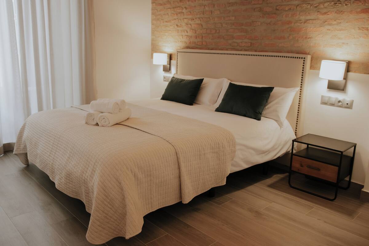 A neatly made bed with a beige quilt, two pillows, and a rolled towel on a wooden floor.