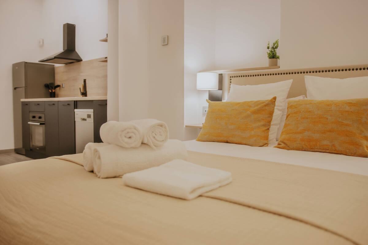 A hotel room with a neatly made bed, a small kitchenette, and a bedside table with a lamp.