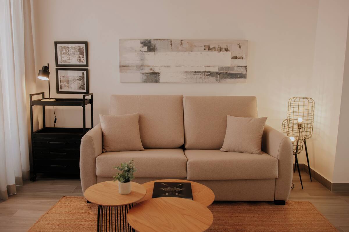 A modern living room with a beige sofa, two side tables, a black desk, and a large abstract painting on the wall.