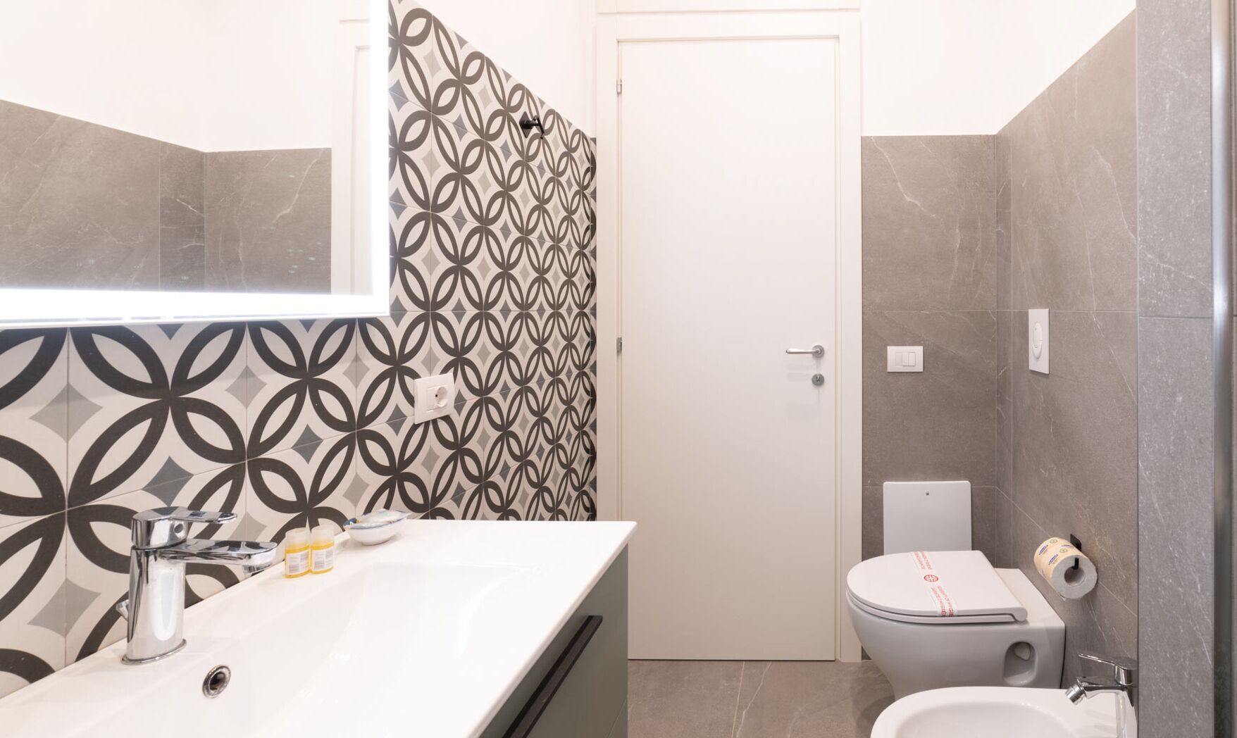 Une salle de bain moderne avec un lavabo, un miroir et des toilettes, le tout de couleur blanche. Les murs sont recouverts de carreaux aux motifs géométriques.