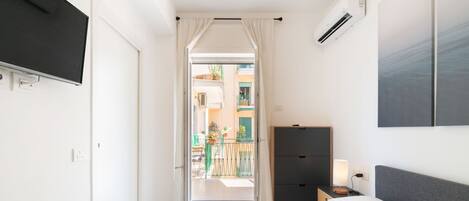 A hotel room with a bed, bedside table, TV, air conditioning unit, and a view of the balcony and outside buildings.
