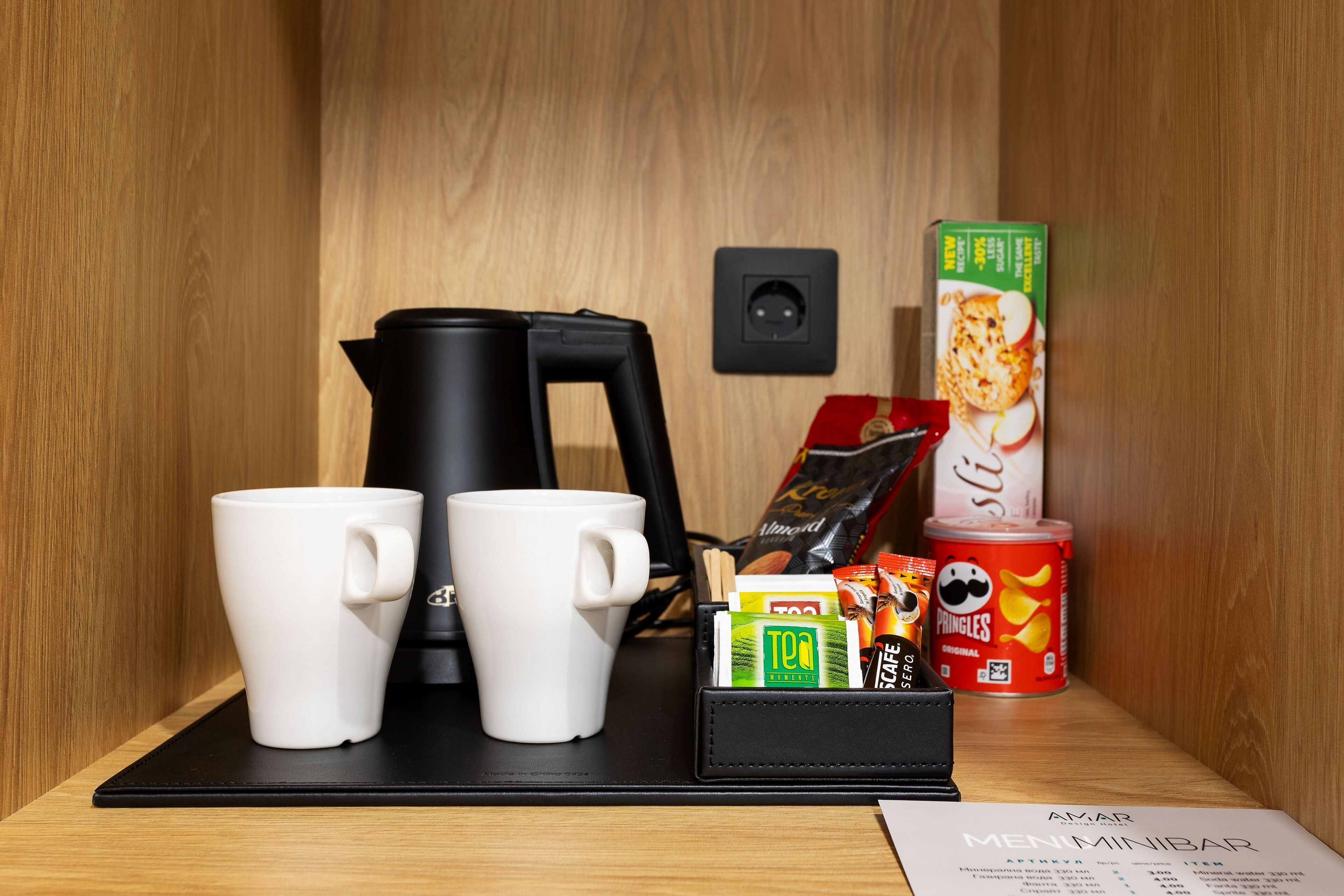 Una estación de café con dos tazas blancas, una cafetera negra, una caja de té, una lata de Pringles y un menú.