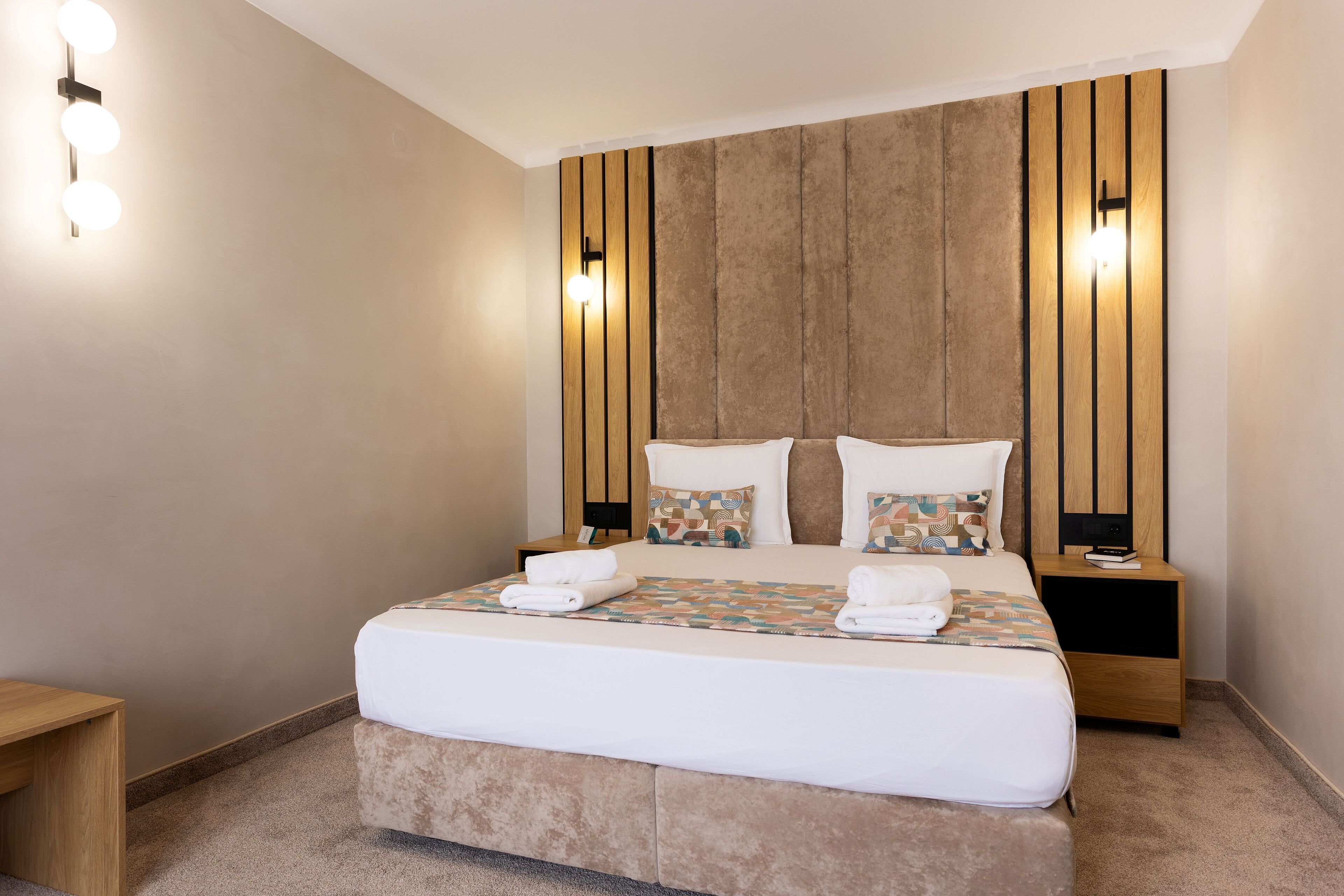 Une chambre d’hôtel avec un grand lit, une tête de lit en bois, des tables de chevet et des lampes fixées au mur.