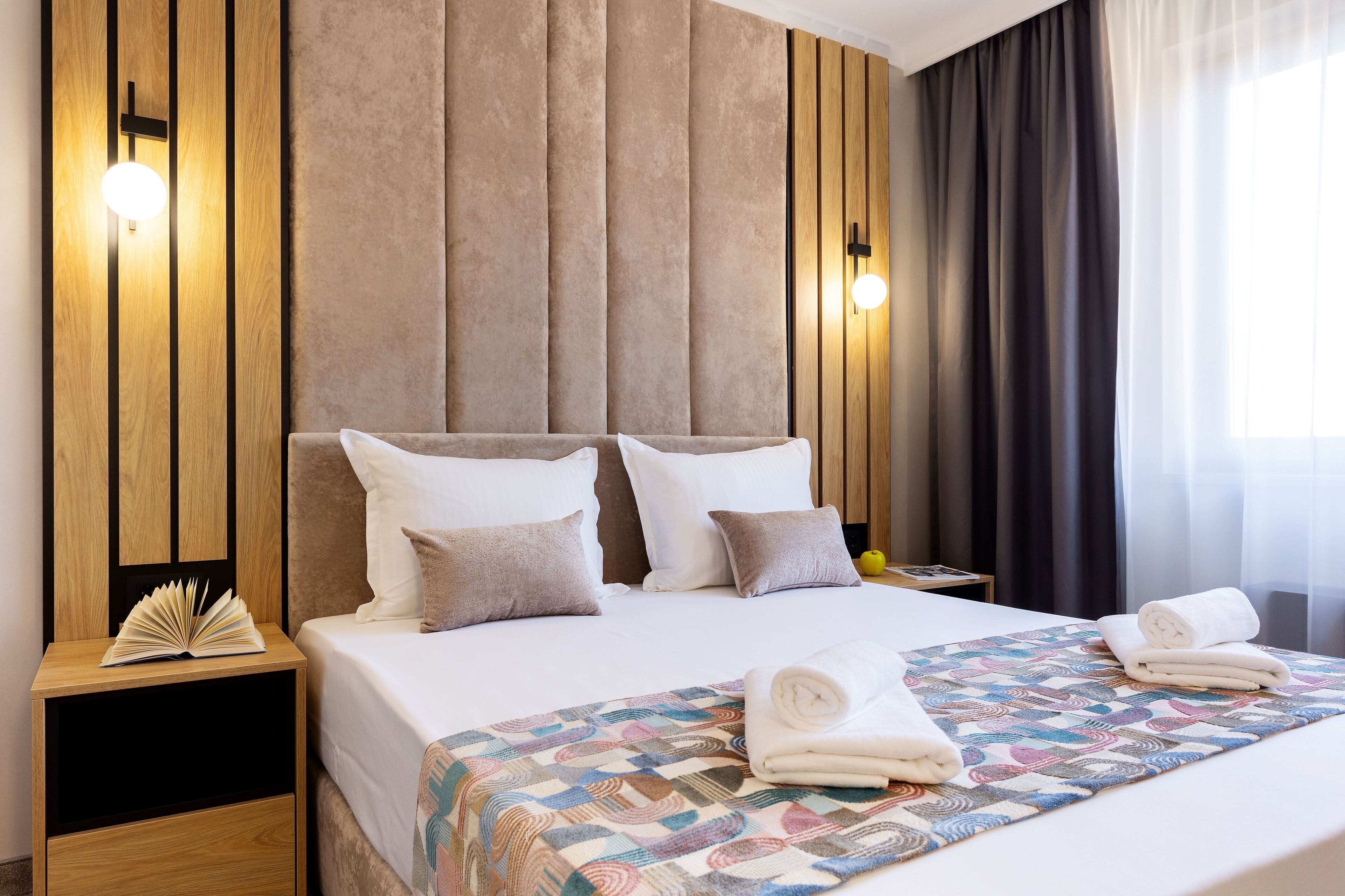 Een net opgemaakt bed met een geborduurde deken en opgerolde handdoeken, een nachtkastje met een boek, en een wand met verticale houten panelen en wandlampen.