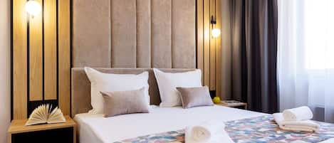 Een net opgemaakt bed met een geborduurde deken en opgerolde handdoeken, een nachtkastje met een boek, en een wand met verticale houten panelen en wandlampen.