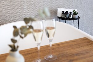 Habitación con una bañera blanca, una mesita con botellas y una planta, y dos copas de champán sobre una bandeja de madera.