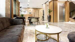 A modern lobby with a plush sofa, a round table with a vase, a reception desk, and a staircase leading to an elevator. - AMAR Design Hotel by HMG (Sofia)