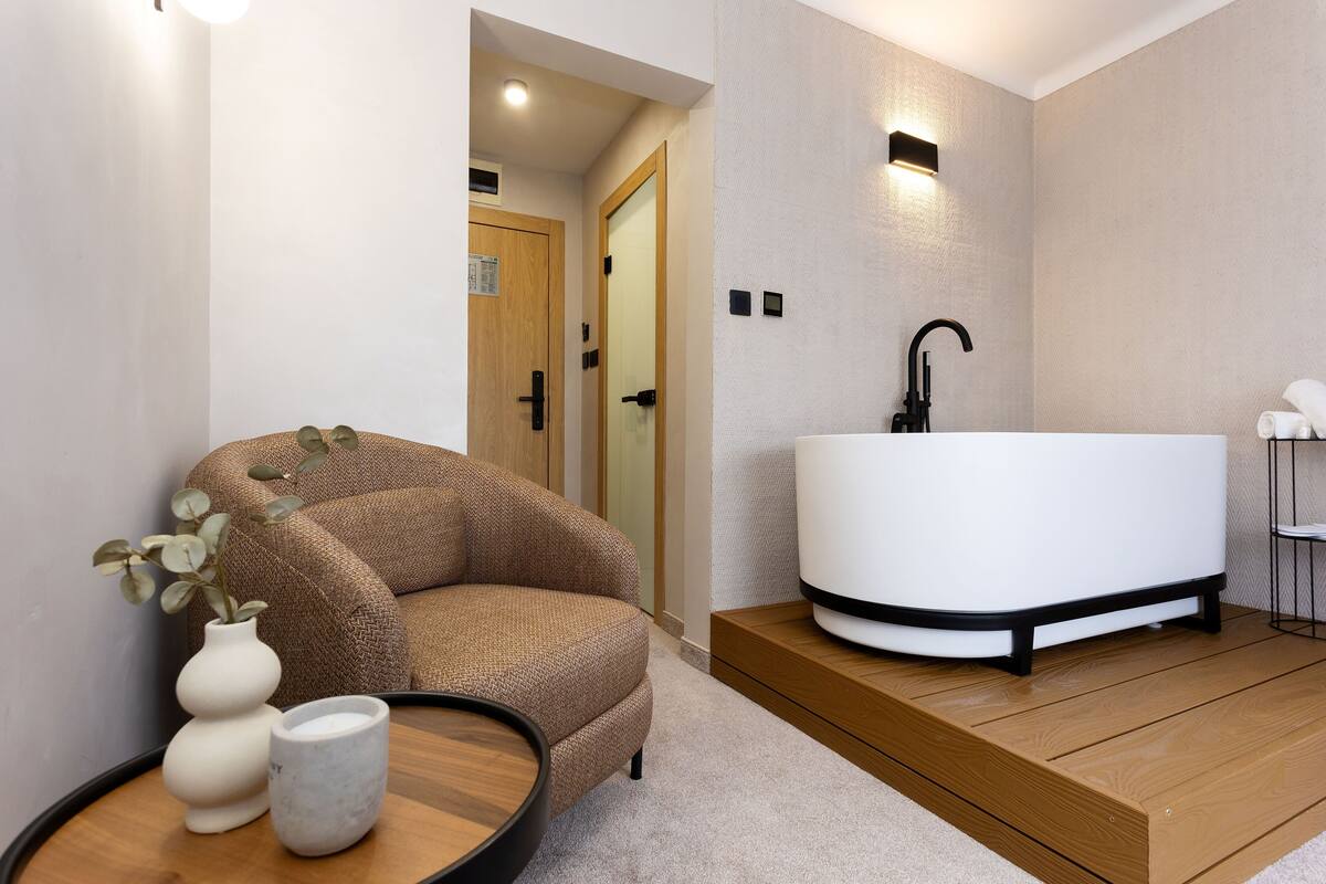 A modern bathroom with a freestanding bathtub, a wooden floor, and a seating area with a plant and a vase.