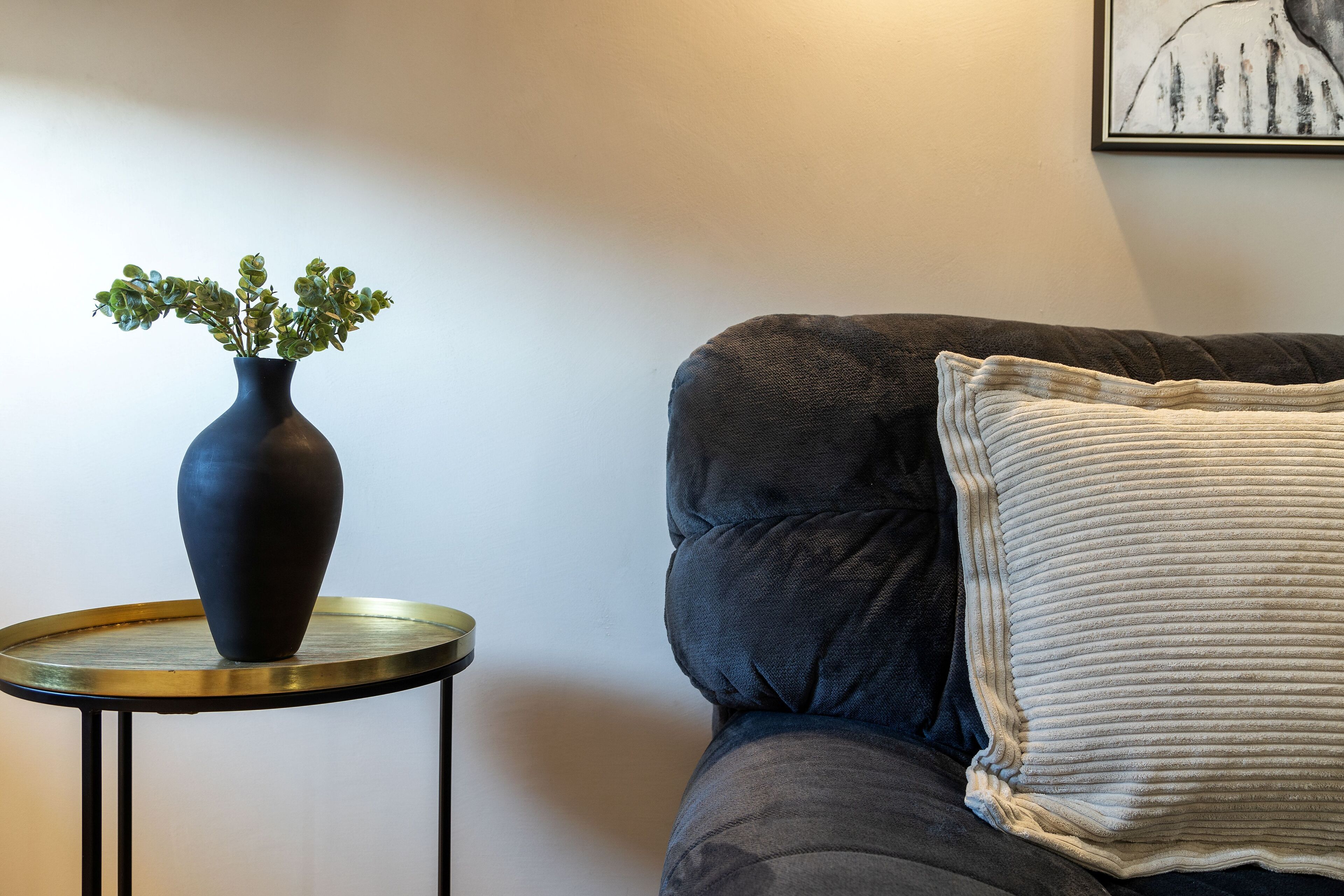 A dark-colored sofa with a textured cushion and a round side table with a dark vase containing green plants.