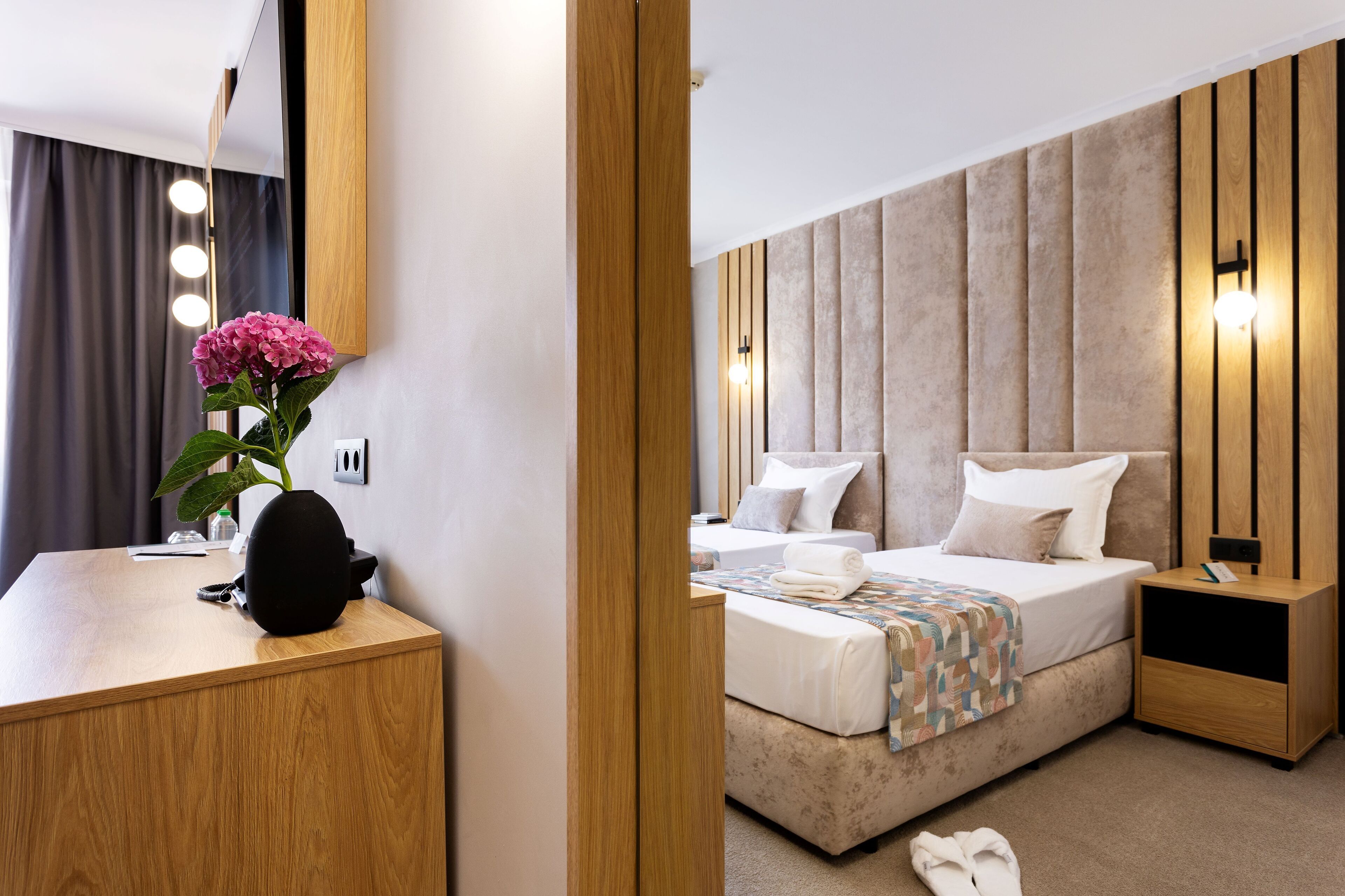 A modern hotel room with a wooden desk, a bed with a beige headboard, and a nightstand.