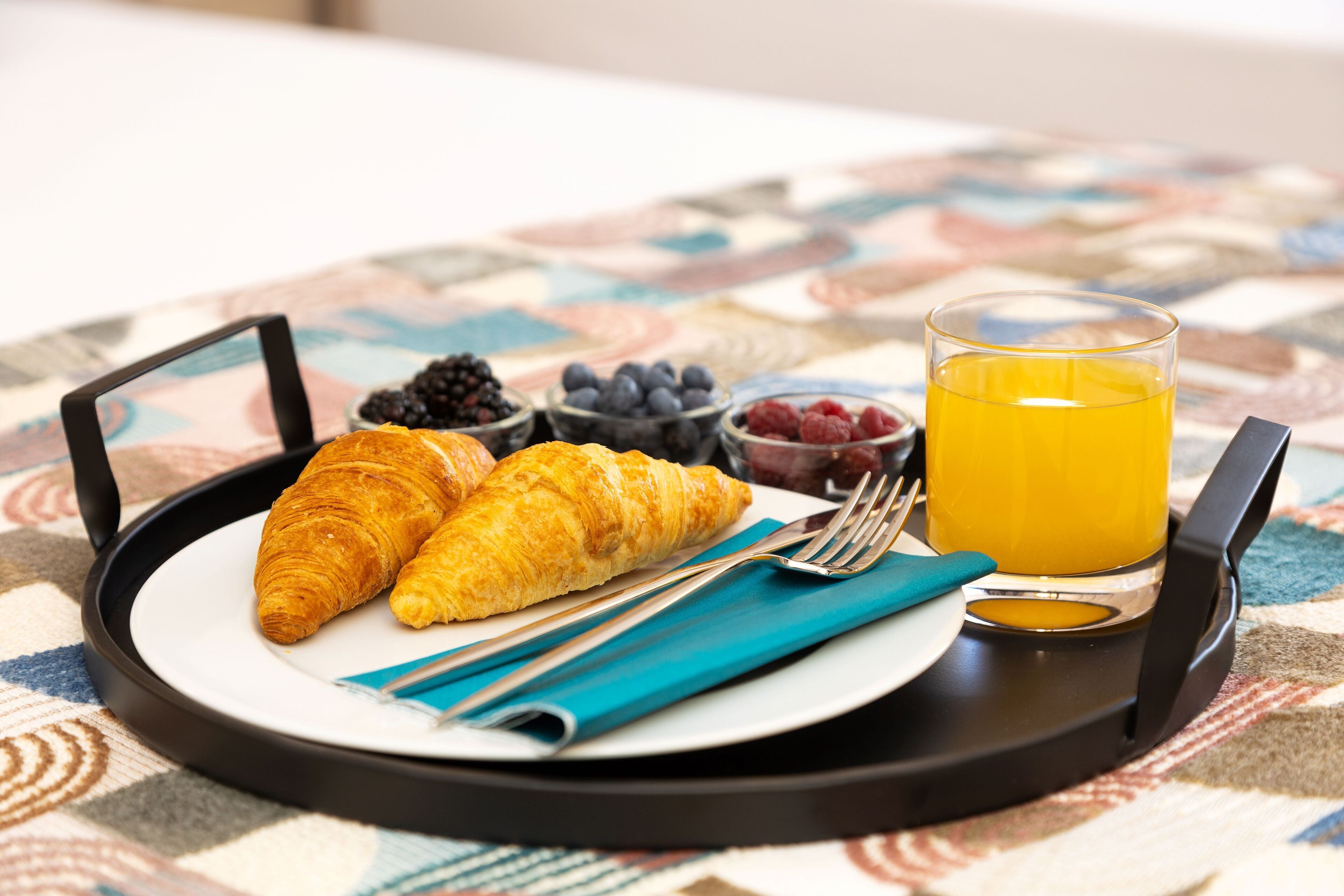 En bakke med morgenmad, blandt andet croissanter, bær og appelsinjuice, placeret på en borddug med et mønster.