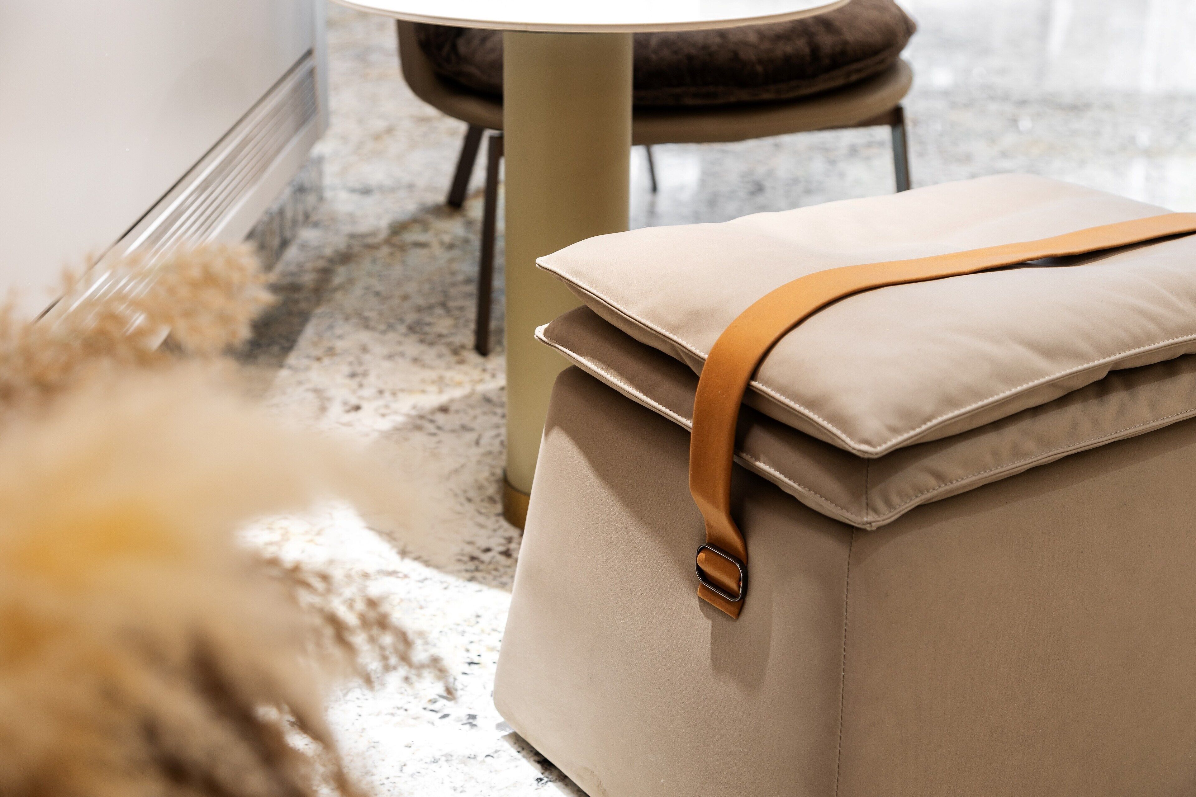 A beige ottoman with a brown strap and a cushion, placed on a speckled floor next to a chair with a brown cushion.