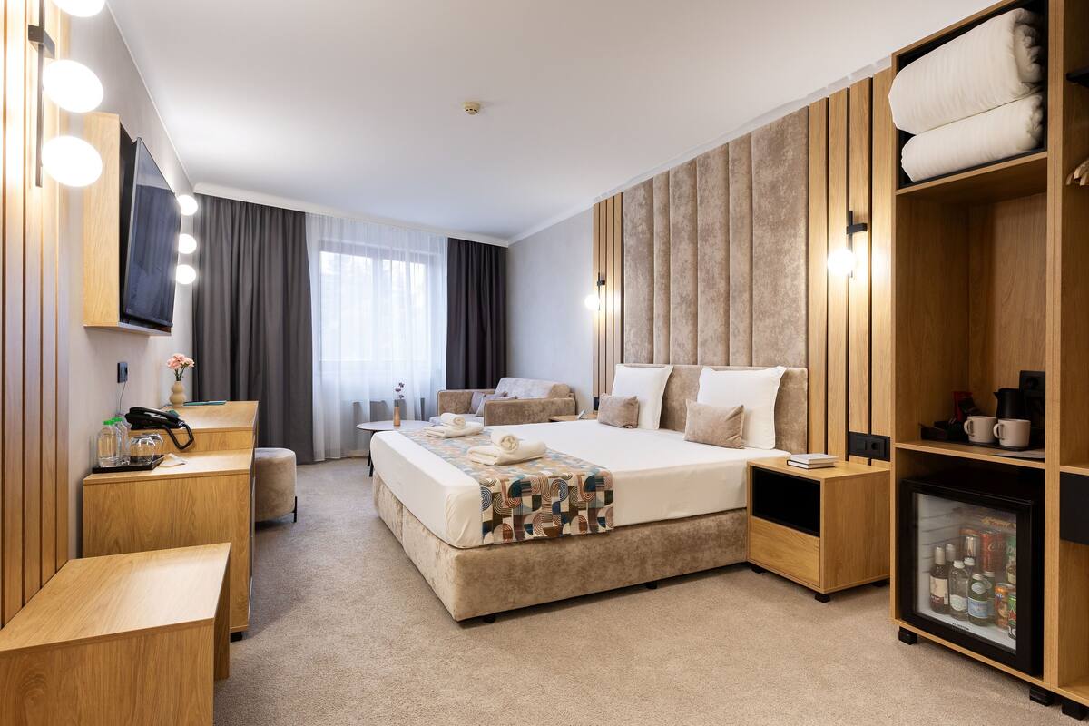 A modern hotel room with a large bed, a seating area, a vanity with a mirror, and a minibar.