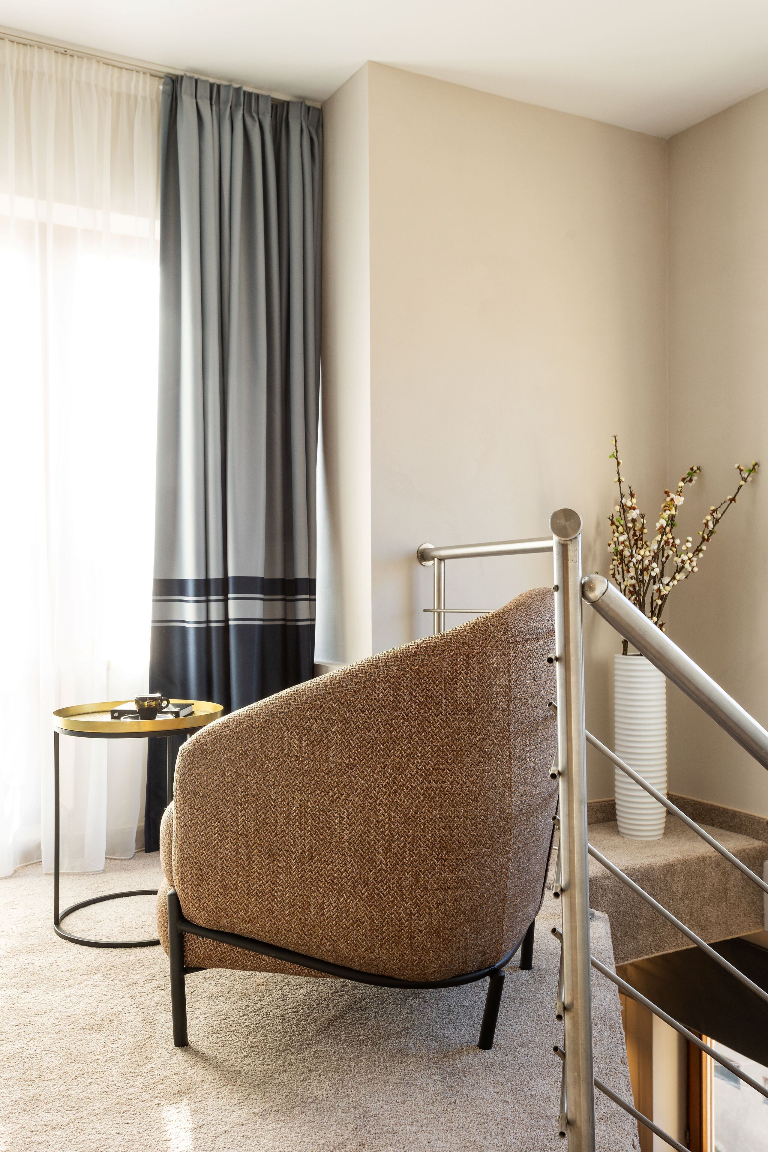 Een moderne woonkamer met een ruime, gestructureerde fauteuil, een klein rond tafeltje met een kopje en een trap met een leuning.