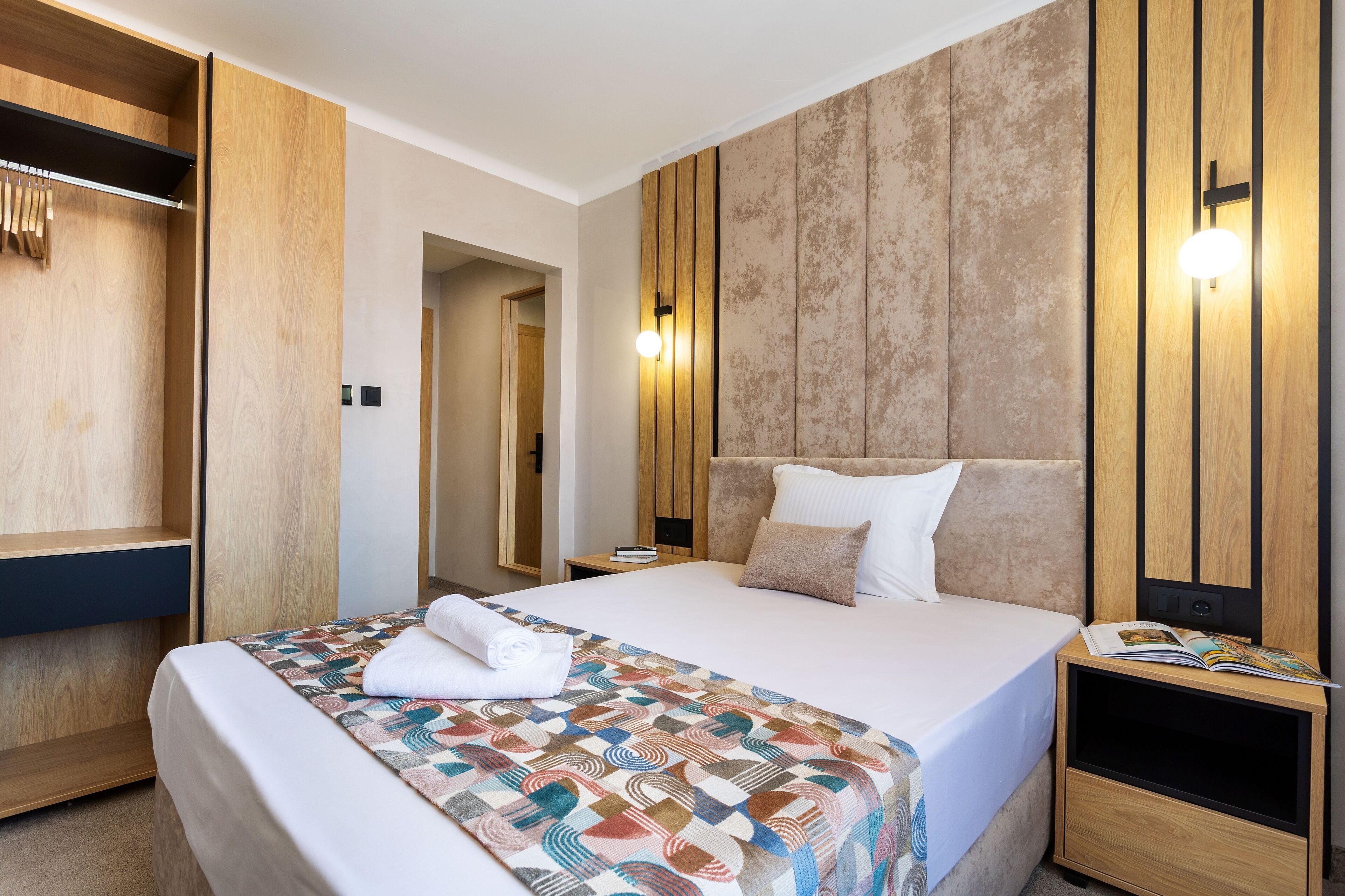 A hotel room with a large bed, wooden wardrobe, and a nightstand with a book and magazine.