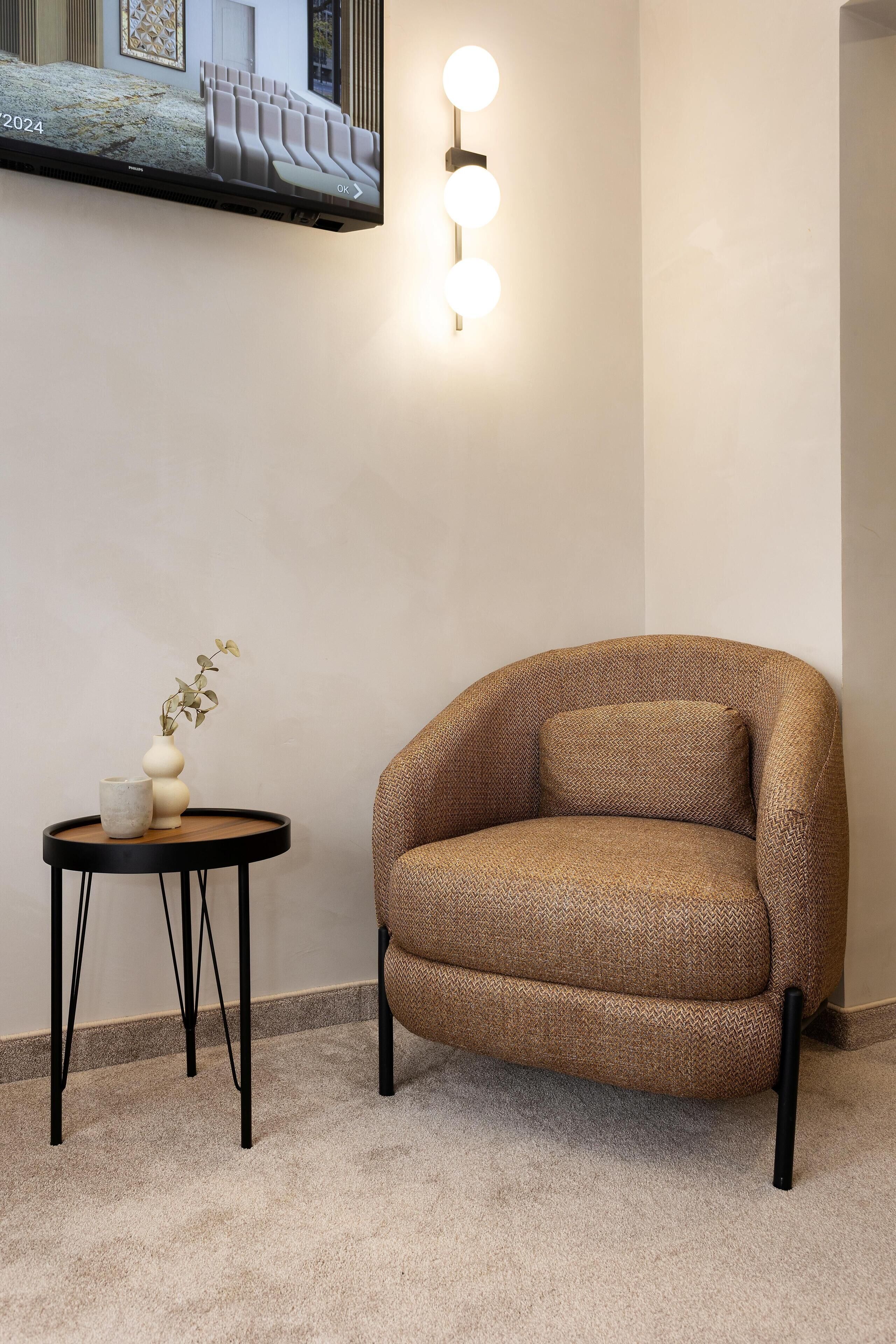 Una habitación moderna con un sillón marrón, una mesita redonda con un jarrón y una planta, y una lámpara fijada a la pared.