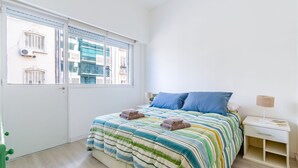 A bedroom with a bed, two pillows, a bedside table with a lamp, and a window with blinds.