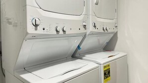 A row of white stacked washer and dryer units with control knobs and a yellow safety sign.